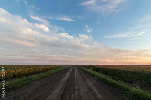 A wide angle landscape shot of an open dirt road among agricultural farming fields with a dramatic sky and orange and blue clouds at sunset