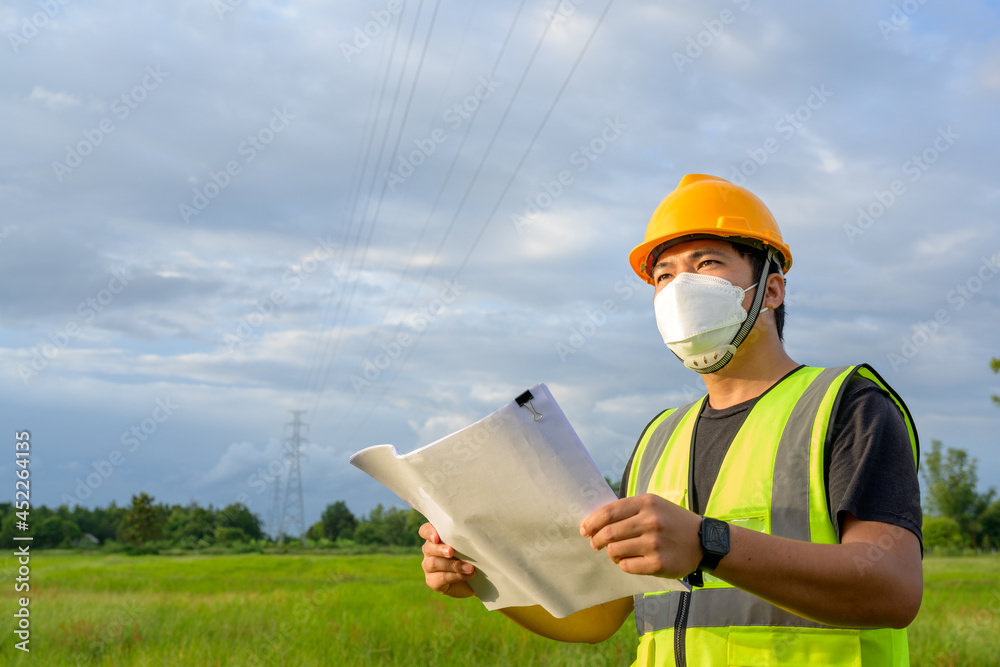 Asian male electrical engineer standing holding a blueprint at the working power plant and plans to inspect high-voltage poles at the power plant