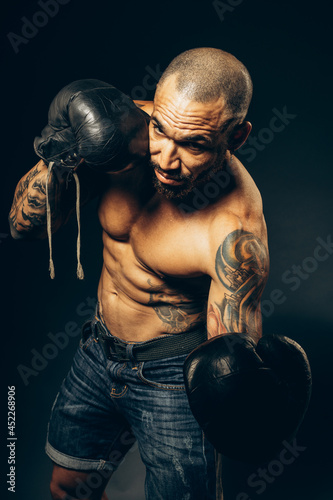 Studio portrait of a muscular boxer with a naked torso on a dark background. The Latin American puncher's attack. A serious fighter performs punches. © SerPak