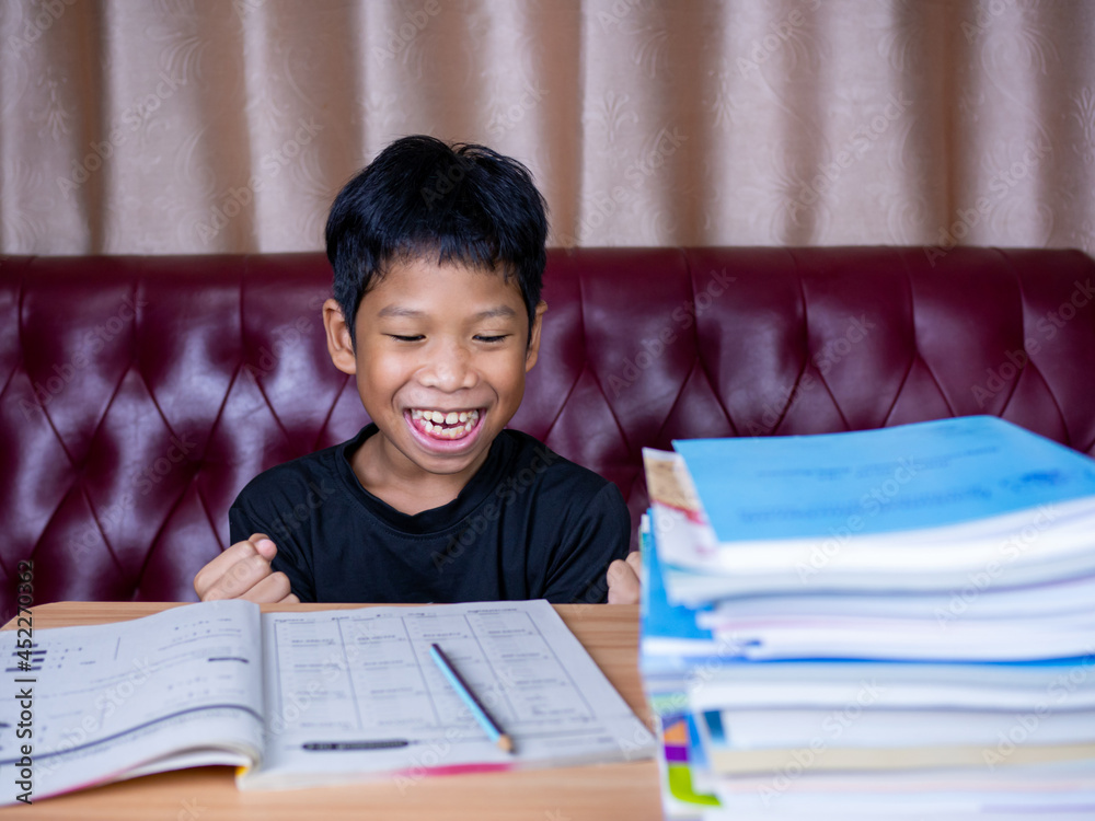 The boy was very happy to finish his homework. He sat on a wooden table and had a stack of books next to it. The background is a red sofa and cream curtains. The boy was very happy to finish his homework. He sat on a wooden table and had a stack of books next to it. The background is a red sofa and cream curtains.