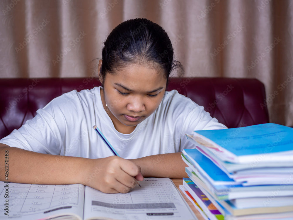 girl doing homework on a wooden table and there was a pile of books next to it The background is a red sofa and cream curtains. girl doing homework on a wooden table and there was a pile of books next to it The background is a red sofa and cream curtains.