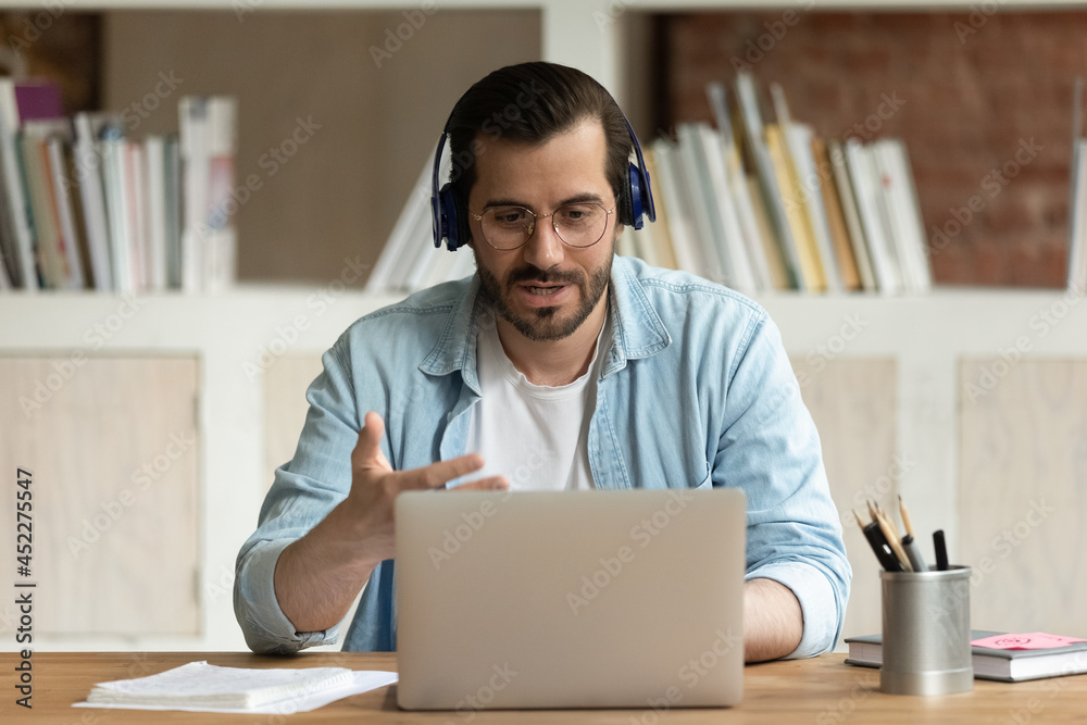 Obraz premium Serious man in glasses headphones sit at workplace desk talks through videoconference, take part in online stream communication use laptop, lead business negotiations remotely. Videocall event concept