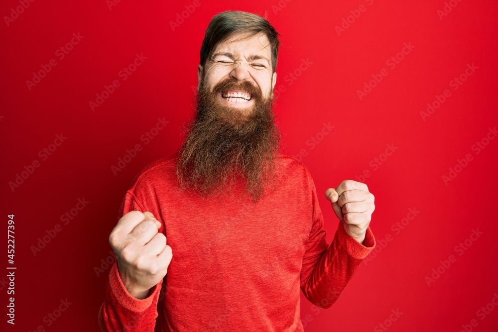 Redhead man with long beard wearing casual clothes celebrating surprised and amazed for success with arms raised and eyes closed Redhead man with long beard wearing casual clothes celebrating surprised and amazed for success with arms raised and eyes closed