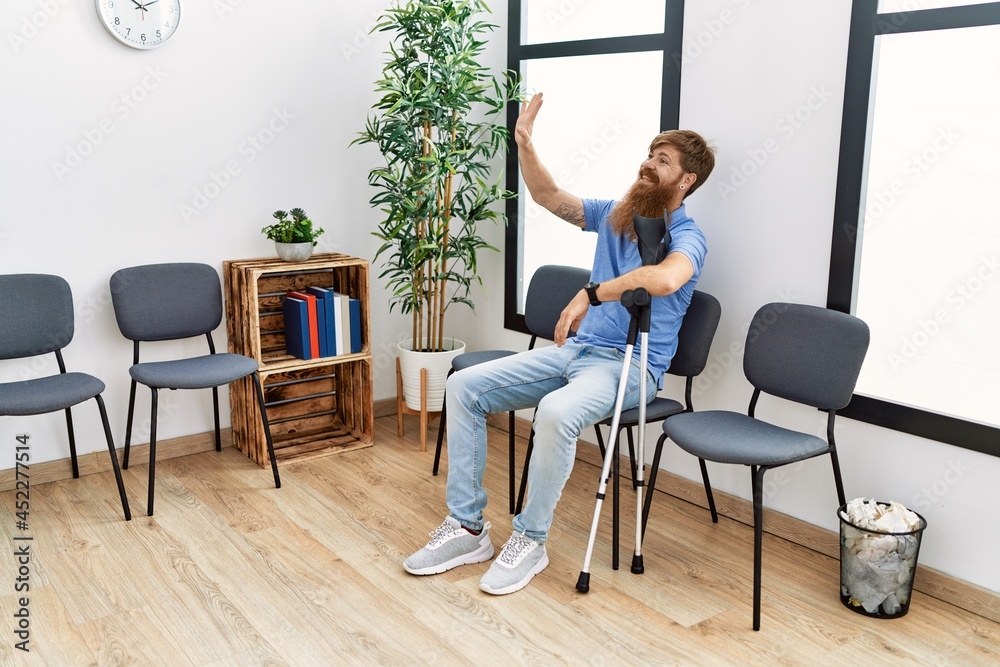 Young redhead man using crutches sitting on the chair at clinic waiting room.