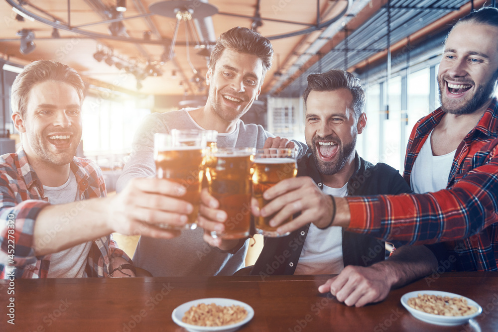 Happy young men in casual clothing toasting each other with beer and laughing while sitting in the pub Happy young men in casual clothing toasting each other with beer and laughing while sitting in the pub