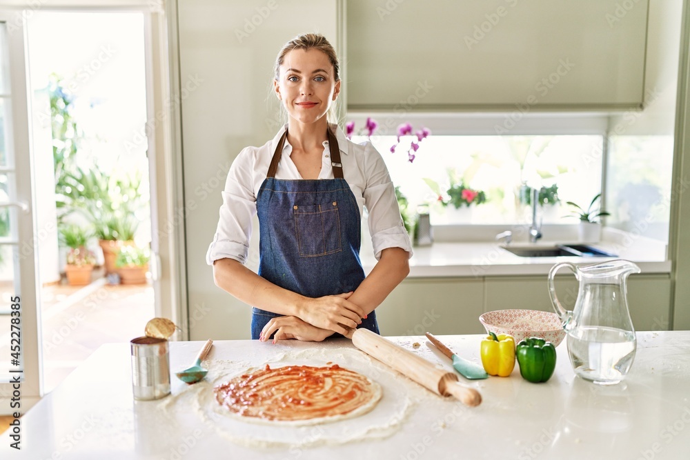 Beautiful blonde woman wearing apron cooking pizza with serious expression on face. simple and natural looking at the camera.