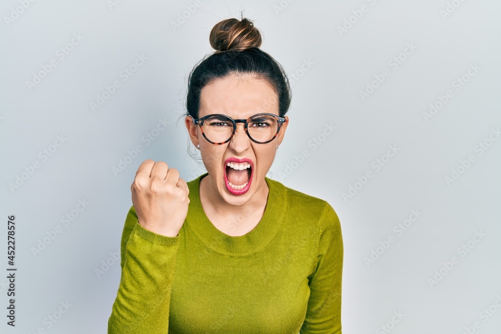 Young hispanic girl wearing casual clothes and glasses angry and mad raising fist frustrated and furious while shouting with anger. rage and aggressive concept.