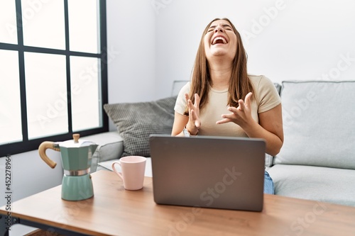 Young brunette woman using laptop at home drinking a cup of coffee celebrating mad and crazy for success with arms raised and closed eyes screaming excited. winner concept
