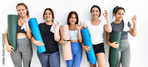 Group of women holding yoga mat standing over isolated background smiling positive doing ok sign with hand and fingers. successful expression.