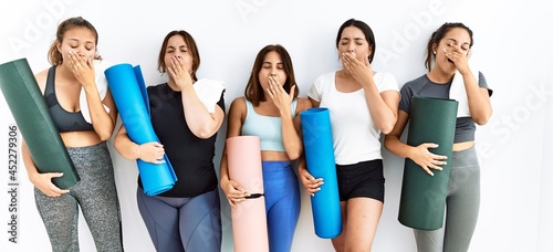 Group of women holding yoga mat standing over isolated background bored yawning tired covering mouth with hand. restless and sleepiness.
