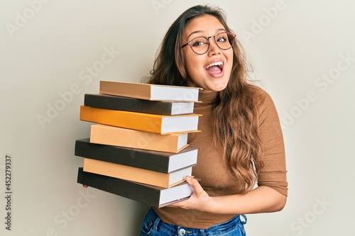 Young hispanic girl holding a pile of books celebrating crazy and amazed for success with open eyes screaming excited.