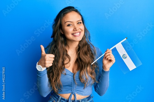 Young hispanic girl holding el salvador flag smiling happy and positive, thumb up doing excellent and approval sign