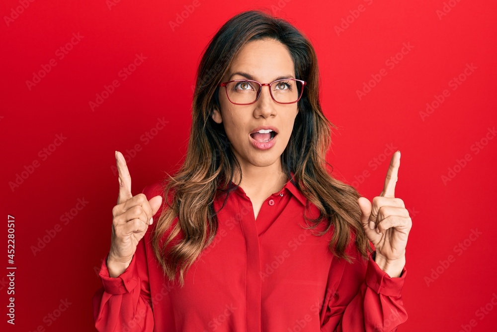 Young latin woman wearing casual clothes and glasses amazed and surprised looking up and pointing with fingers and raised arms. Young latin woman wearing casual clothes and glasses amazed and surprised looking up and pointing with fingers and raised arms.