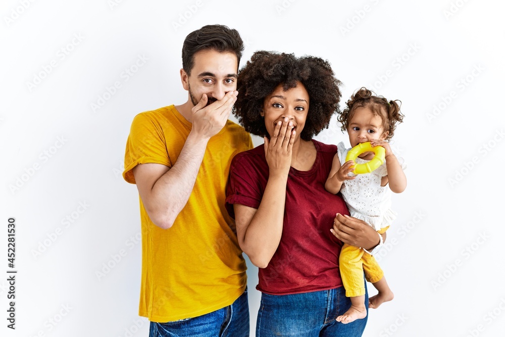 Interracial young family of black mother and hispanic father with daughter laughing and embarrassed giggle covering mouth with hands, gossip and scandal concept Interracial young family of black mother and hispanic father with daughter laughing and embarrassed giggle covering mouth with hands, gossip and scandal concept