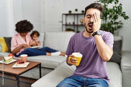 Hispanic father of interracial family drinking a cup coffee yawning tired covering half face  eye and mouth with hand. face hurts in pain.