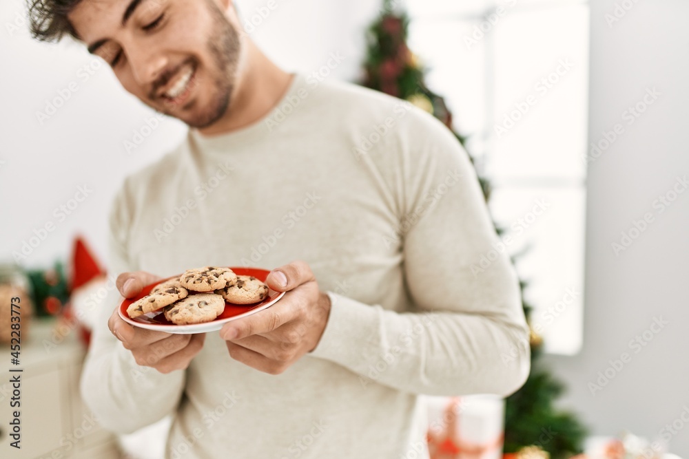 Young hispanic man wearing christmas hat holding cookies at home.