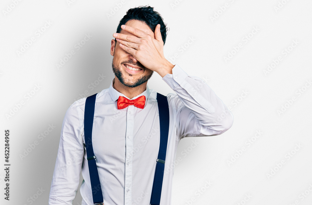 Hispanic man with beard wearing hipster look with bow tie and suspenders smiling and laughing with hand on face covering eyes for surprise. blind concept. Hispanic man with beard wearing hipster look with bow tie and suspenders smiling and laughing with hand on face covering eyes for surprise. blind concept.
