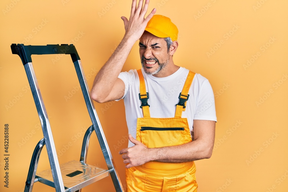 Handsome middle age man with grey hair holding ladder surprised with hand on head for mistake, remember error. forgot, bad memory concept.