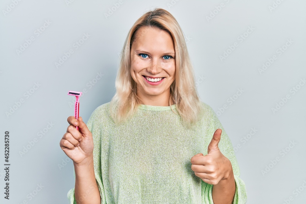 Beautiful caucasian blonde woman holding razor smiling happy and positive, thumb up doing excellent and approval sign Beautiful caucasian blonde woman holding razor smiling happy and positive, thumb up doing excellent and approval sign