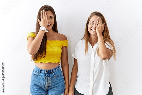 Mother and daughter together standing together over isolated background covering one eye with hand, confident smile on face and surprise emotion.