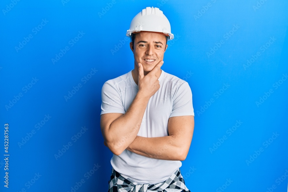 Handsome young man wearing builder uniform and hardhat looking confident at the camera smiling with crossed arms and hand raised on chin. thinking positive. Handsome young man wearing builder uniform and hardhat looking confident at the camera smiling with crossed arms and hand raised on chin. thinking positive.