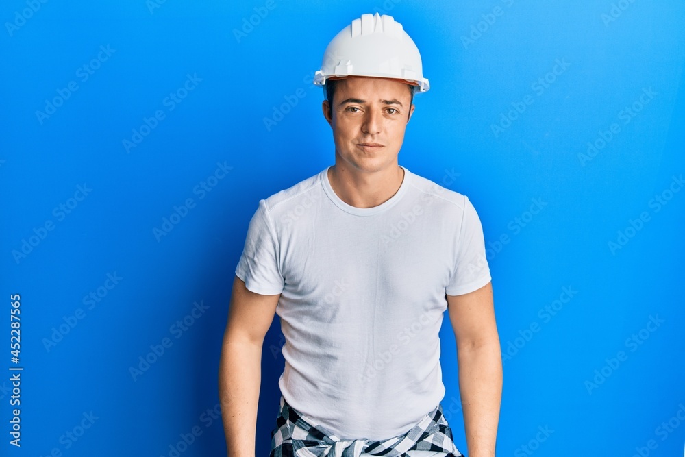 Handsome young man wearing builder uniform and hardhat relaxed with serious expression on face. simple and natural looking at the camera.