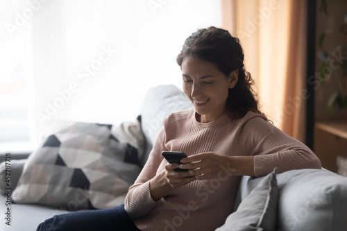 Happy beautiful Latina woman relaxing on comfortable couch holding smartphone in hands chatting in social media networks, watching funny videos online, use new cool freeware mobile application on cell