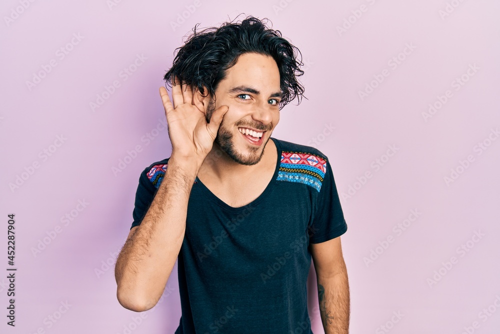 Handsome hispanic man wearing casual t shirt smiling with hand over ear listening and hearing to rumor or gossip. deafness concept. Handsome hispanic man wearing casual t shirt smiling with hand over ear listening and hearing to rumor or gossip. deafness concept.
