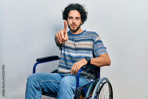 Handsome hispanic man sitting on wheelchair pointing with finger up and angry expression, showing no gesture