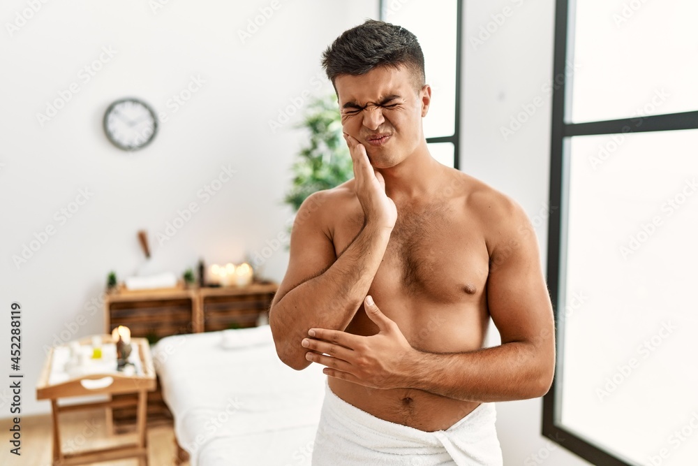 Obraz premium Young hispanic man standing shirtless at spa center touching mouth with hand with painful expression because of toothache or dental illness on teeth. dentist