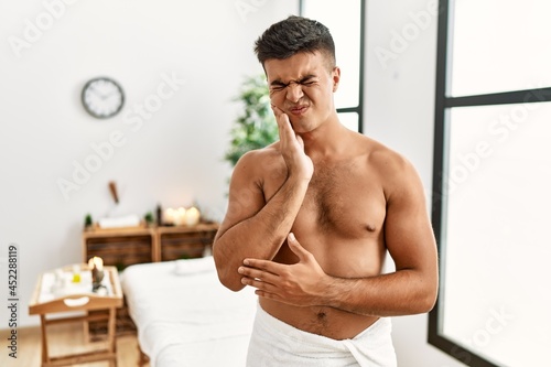 Young hispanic man standing shirtless at spa center touching mouth with hand with painful expression because of toothache or dental illness on teeth. dentist