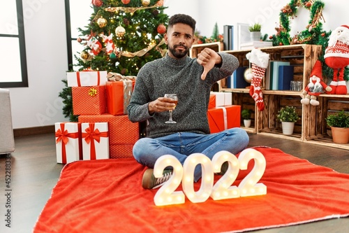 Young hispanic man with beard sitting by christmas tree celebrating 2022 new year with angry face, negative sign showing dislike with thumbs down, rejection concept