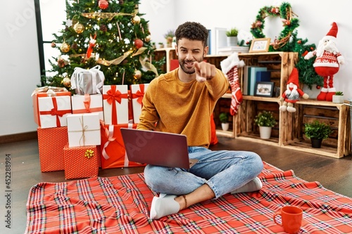 Arab young man using laptop sitting by christmas tree pointing to you and the camera with fingers, smiling positive and cheerful