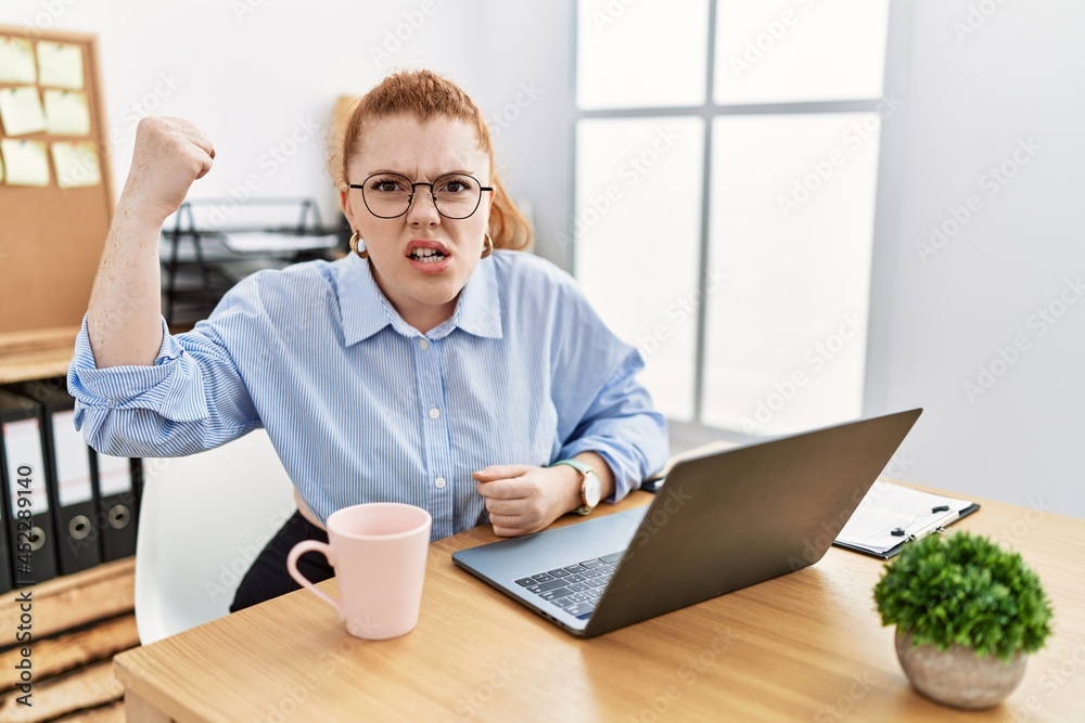 Young redhead woman working at the office using computer laptop angry and mad raising fist frustrated and furious while shouting with anger. rage and aggressive concept.