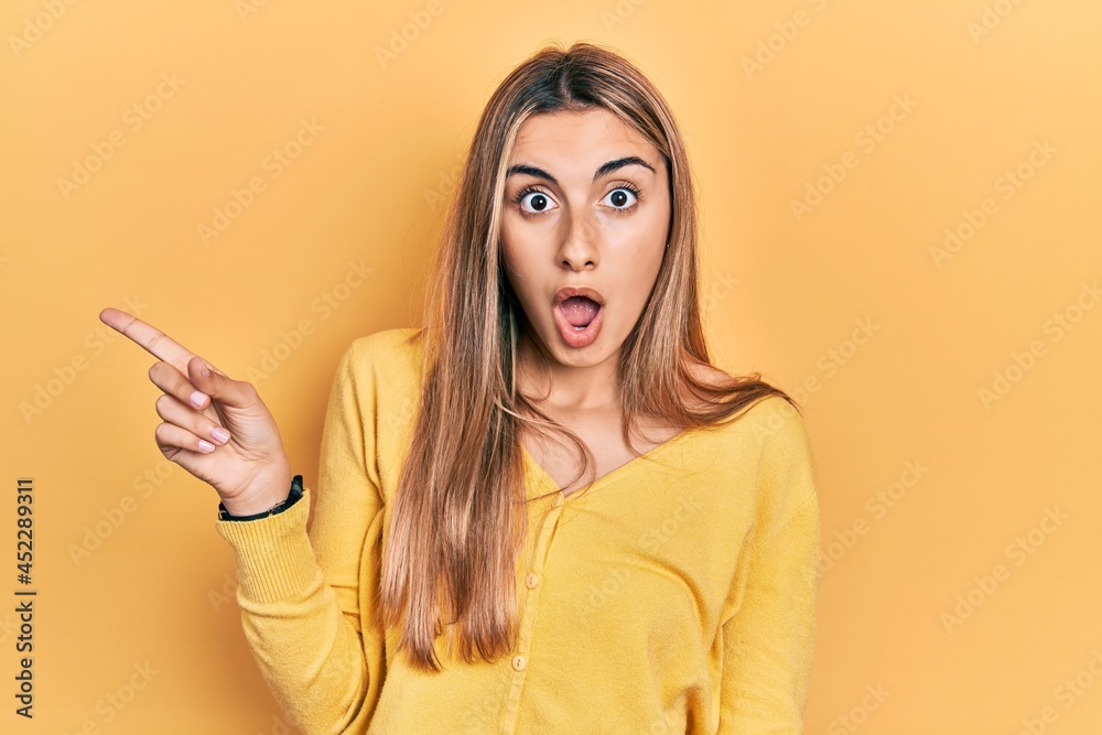 Beautiful hispanic woman wearing casual yellow sweater surprised pointing with finger to the side, open mouth amazed expression. Beautiful hispanic woman wearing casual yellow sweater surprised pointing with finger to the side, open mouth amazed expression.