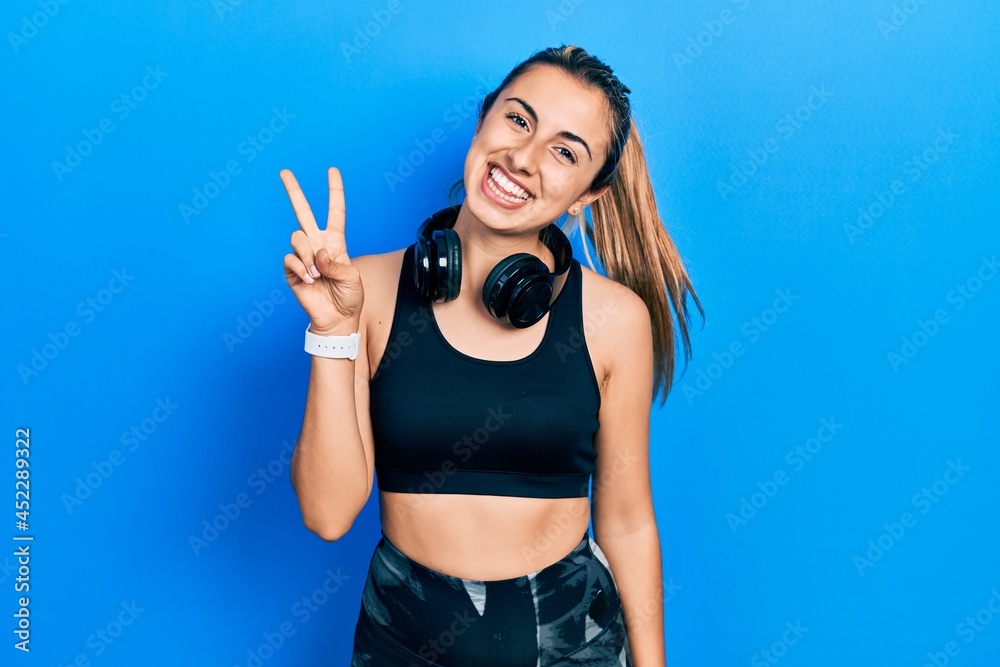 Beautiful hispanic woman wearing gym clothes and using headphones showing and pointing up with fingers number two while smiling confident and happy.
