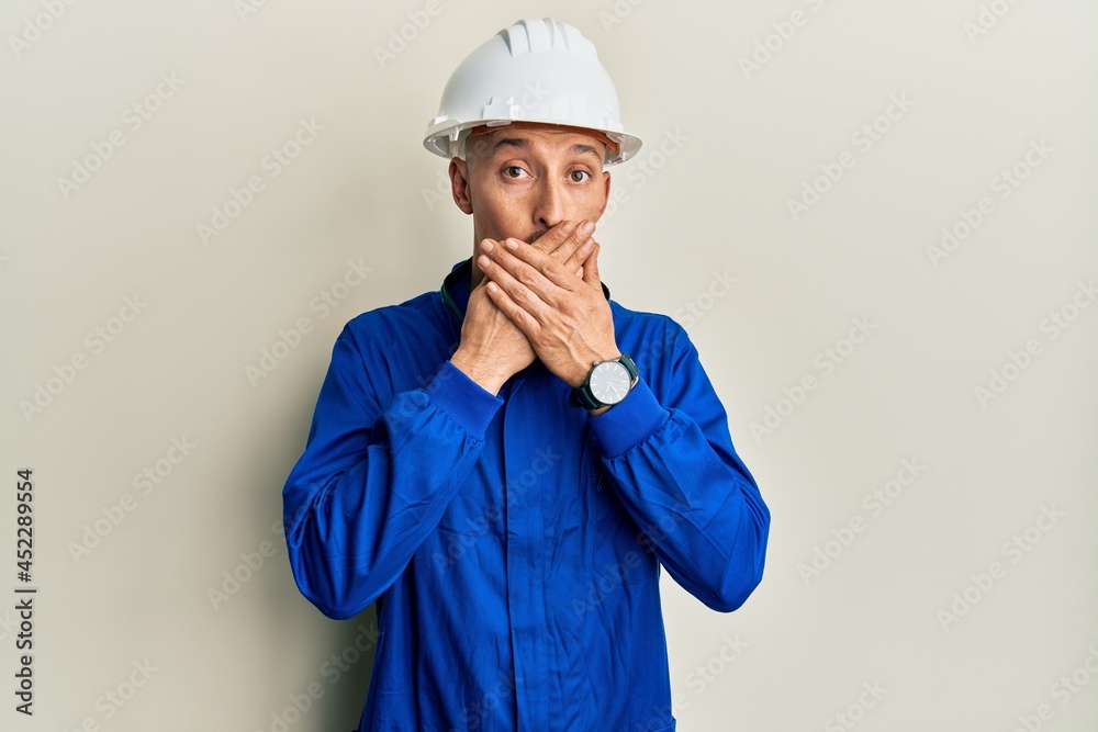 Bald man with beard wearing builder jumpsuit uniform and hardhat shocked covering mouth with hands for mistake. secret concept.
