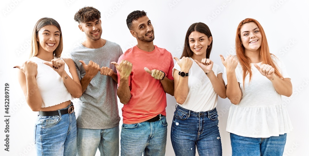 Obraz premium Group of young friends standing together over isolated background pointing to the back behind with hand and thumbs up, smiling confident