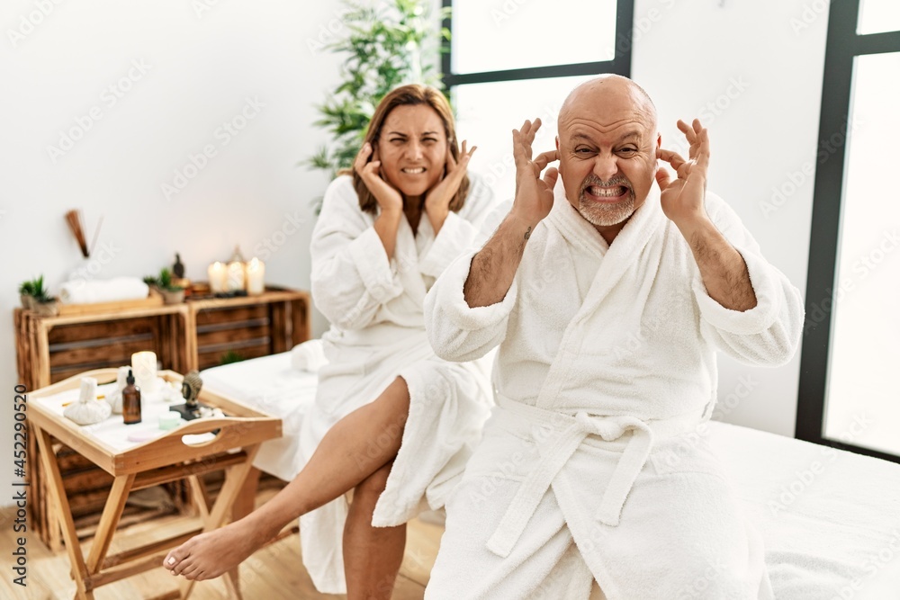Middle age hispanic couple wearing bathrobe at wellness spa covering ears with fingers with annoyed expression for the noise of loud music. deaf concept.