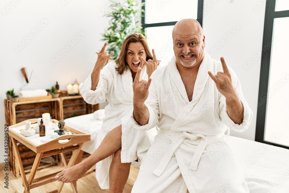 Middle age hispanic couple wearing bathrobe at wellness spa shouting with crazy expression doing rock symbol with hands up. music star. heavy concept. Middle age hispanic couple wearing bathrobe at wellness spa shouting with crazy expression doing rock symbol with hands up. music star. heavy concept.