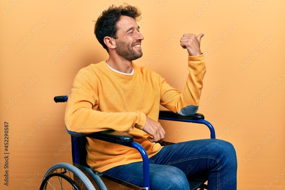 Handsome man with beard sitting on wheelchair smiling with happy face looking and pointing to the side with thumb up. Handsome man with beard sitting on wheelchair smiling with happy face looking and pointing to the side with thumb up.