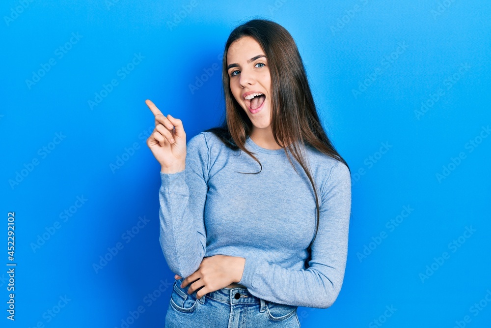 Young brunette teenager wearing casual sweater with a big smile on face, pointing with hand and finger to the side looking at the camera. Young brunette teenager wearing casual sweater with a big smile on face, pointing with hand and finger to the side looking at the camera.