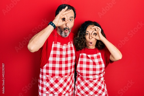 Middle age couple of hispanic woman and man wearing cook apron doing ok gesture shocked with surprised face, eye looking through fingers. unbelieving expression.
