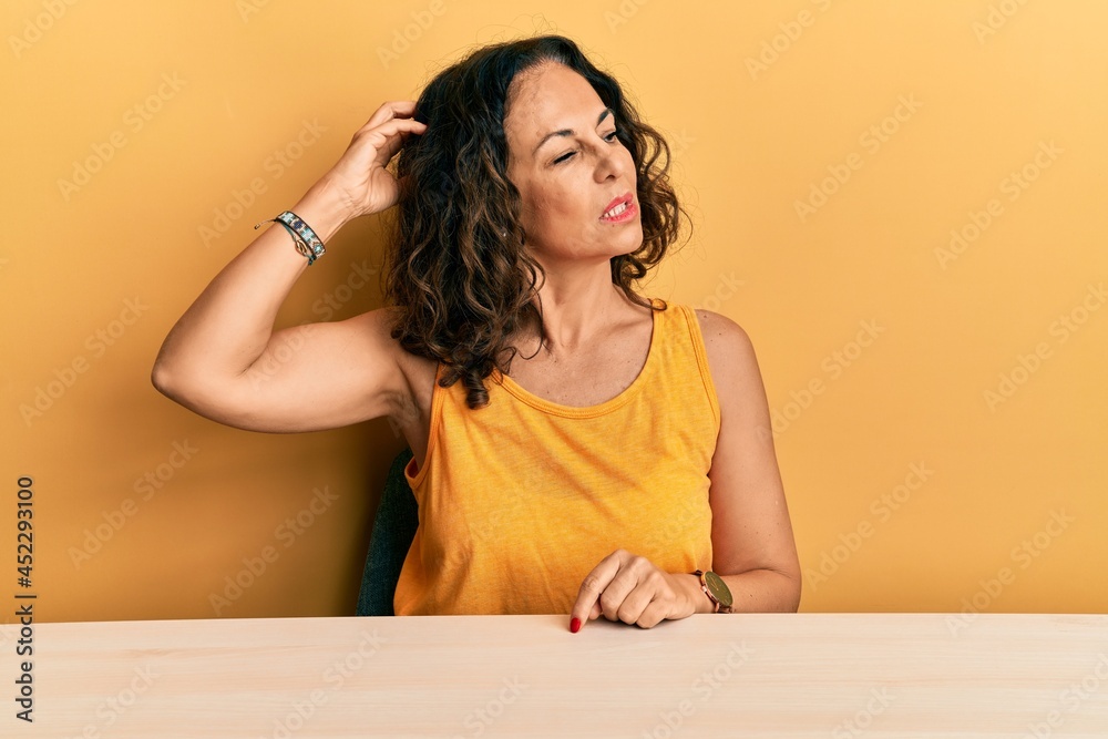 Beautiful middle age woman wearing casual clothes sitting on the table confuse and wondering about question. uncertain with doubt, thinking with hand on head. pensive concept. Beautiful middle age woman wearing casual clothes sitting on the table confuse and wondering about question. uncertain with doubt, thinking with hand on head. pensive concept.