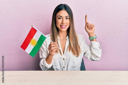 Young hispanic woman holding kurdistan flag sitting on the table smiling with an idea or question pointing finger with happy face, number one photo