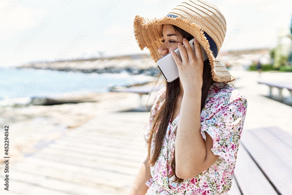 Young latin girl wearing summer talking on the smartphone sitting on the bench hat at the beach.