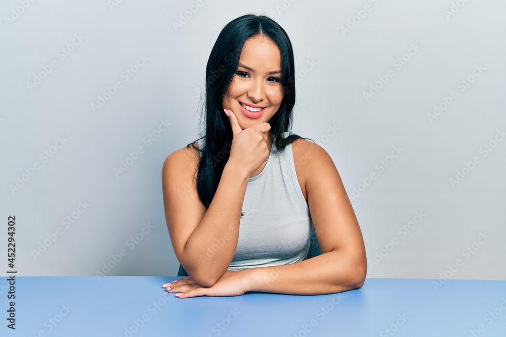 Beautiful hispanic woman with nose piercing sitting on the table looking confident at the camera smiling with crossed arms and hand raised on chin. thinking positive. Beautiful hispanic woman with nose piercing sitting on the table looking confident at the camera smiling with crossed arms and hand raised on chin. thinking positive.
