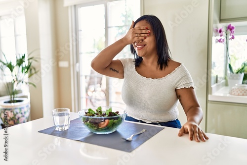 Young hispanic woman eating healthy salad at home smiling and laughing with hand on face covering eyes for surprise. blind concept.