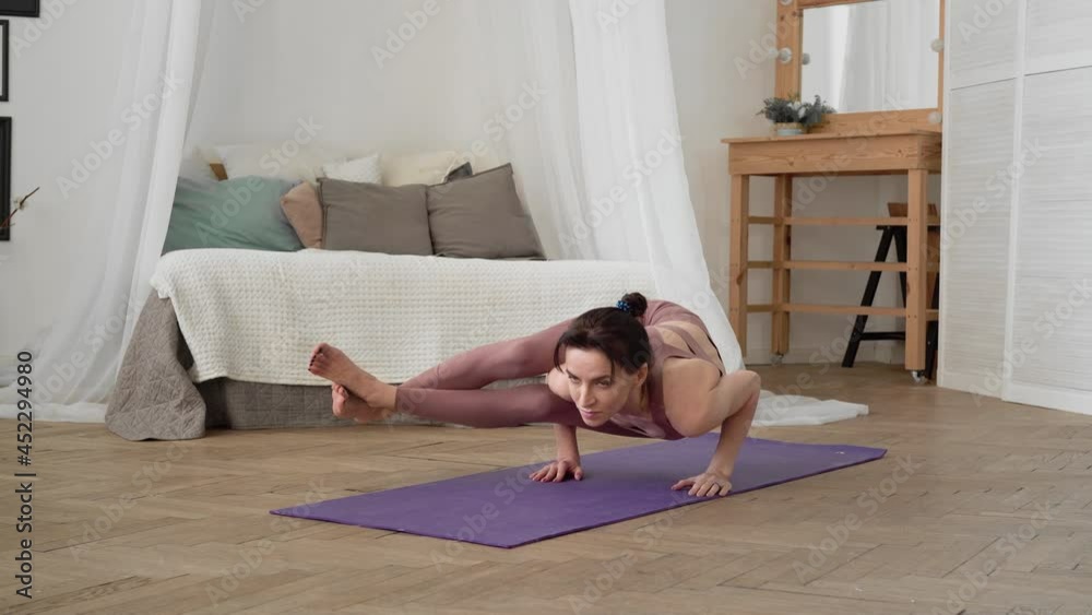 Slender, flexible, caucasian middle-aged woman with black hair in beige tracksuit performs standing asanas one-legged king pigeon pose on purple yoga mat in bright room with bed, a mirror, a window Slender, flexible, caucasian middle-aged woman with black hair in beige tracksuit performs standing asanas one-legged king pigeon pose on purple yoga mat in bright room with bed, a mirror, a window