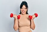 Young hispanic woman wearing sportswear using dumbbells winking looking at the camera with sexy expression, cheerful and happy face.